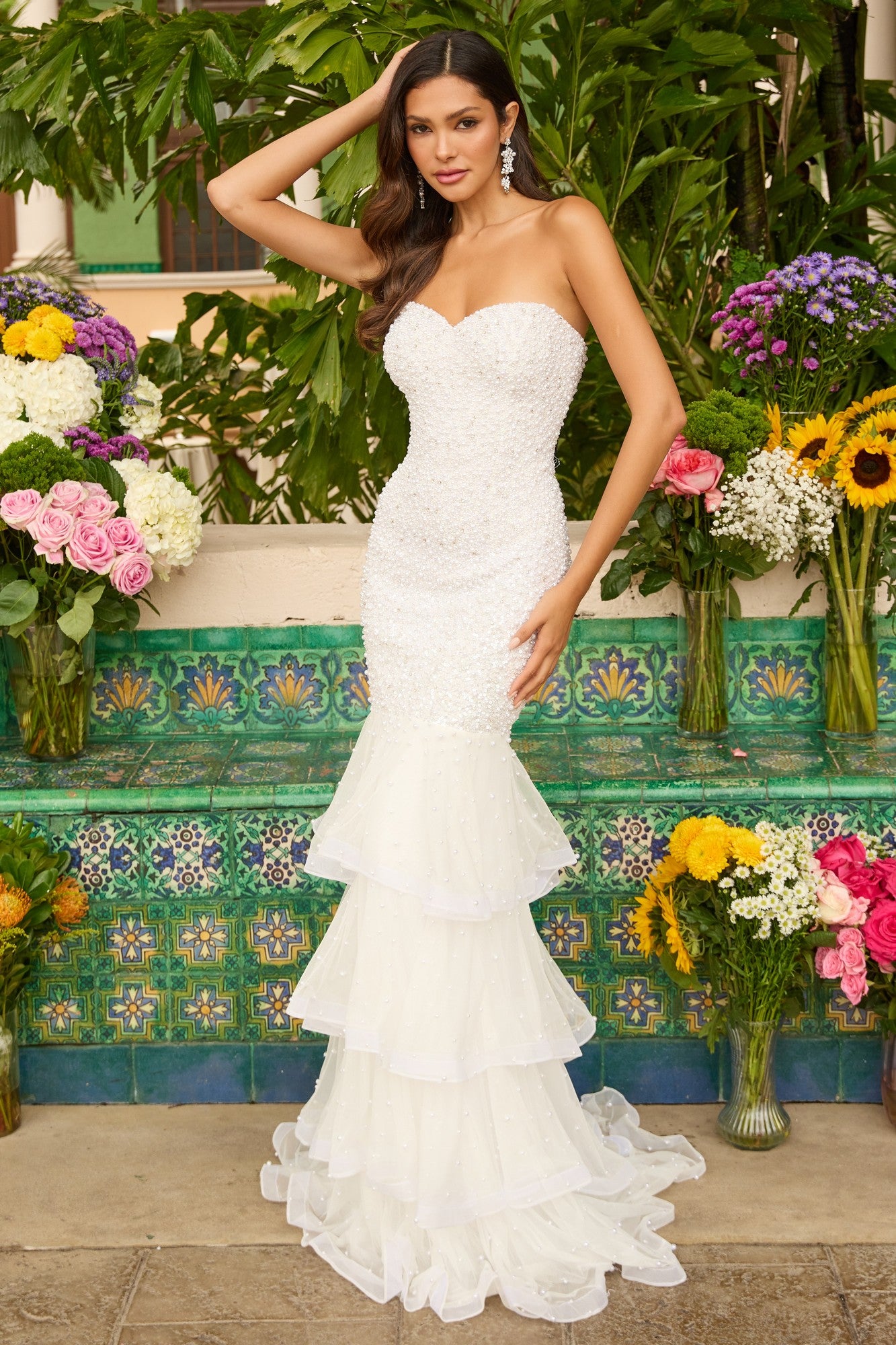 A woman in the ivory Ava Presley 47251 long prom dress, featuring a strapless mermaid silhouette and tiered ruffle layers, stands before colorful tiles and vibrant floral arrangements.